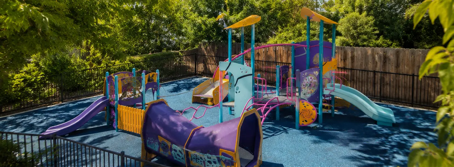 Wide view of Creekside’s shaded playground with slides, climbing equipment, and a soft blue play surface